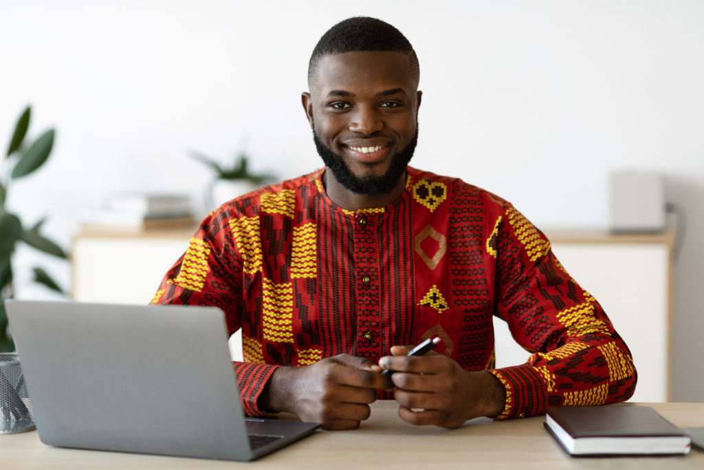 African Freelancer Guy In Traditional Costume Sitting At Desk At Home Office